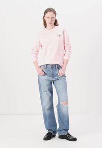 Felpa rosa chiaro a maniche lunghe con una piccola grafica sul petto, abbinata a jeans chiari strappati e scarpe nere.