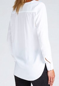 Blusa bianca a maniche lunghe con orlo arrotondato, dettaglio della piega posteriore e polsini con spacco laterale, realizzata in tessuto morbido.