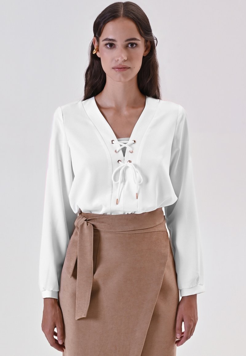 Una donna con lunghi capelli scuri indossa una blusa bianca con lacci a maniche lunghe e una gonna avvolgente beige con cintura annodata, in piedi su uno sfondo grigio semplice.