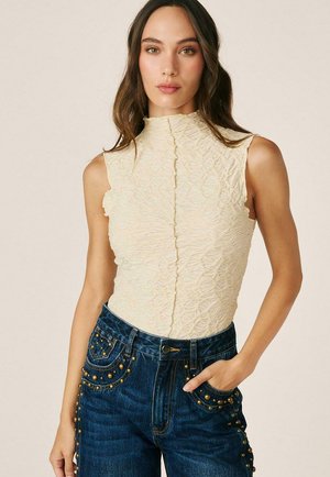Mujer joven con un top sin mangas de cuello alto texturizado en color crema y vaqueros azul oscuro con detalles de tachuelas doradas, de pie con una mano en el bolsillo.