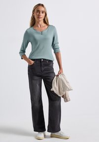 Helle blaue langärmelige Bluse mit rundem Ausschnitt, kombiniert mit dunkelgrauen, weitgeschnittenen Jeans und weißen Sneakers, während sie eine beigefarbene Jacke hält.