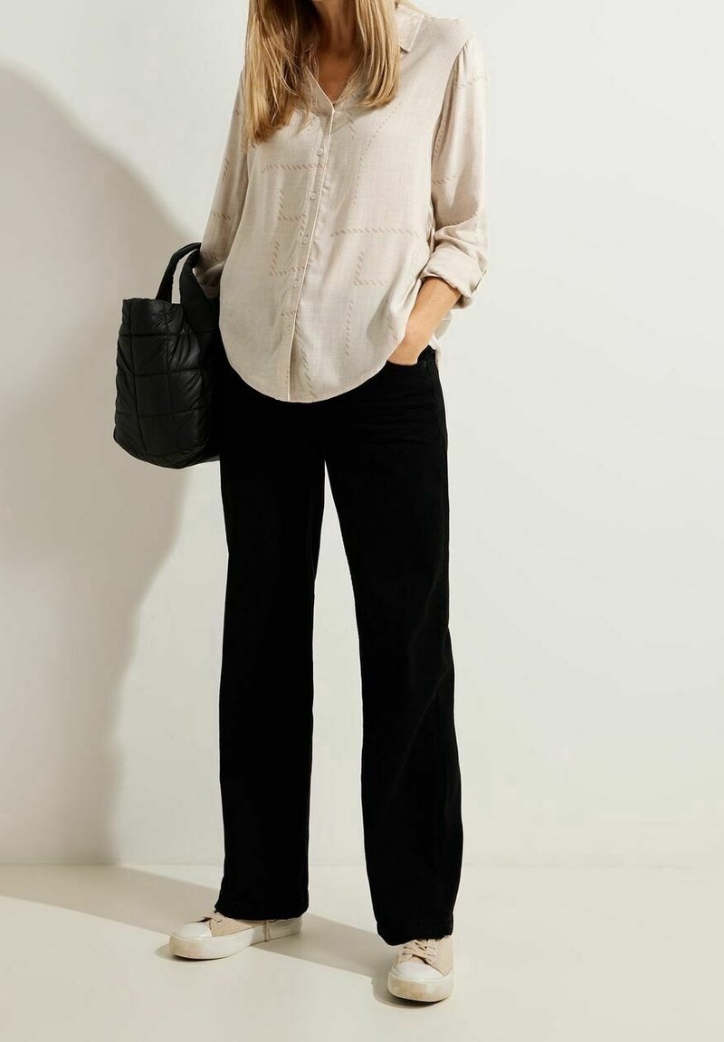 Femme portant une chemise beige boutonnée, un pantalon large noir, des baskets blanches, tenant un sac cabas noir matelassé, debout contre un mur uni.