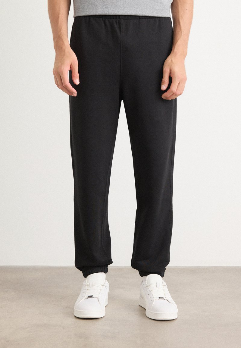 Sorte joggerbukser lavet af blødt stof, med elastisk talje og manchetter. Sammen med hvide sneakers på et neutralt gulv.