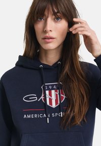 Navy hoodie met een groot logo met rode en witte strepen, trekkoordsluiting en een zak aan de voorkant. Zachte stof met een casual ontwerp.