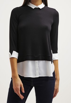 Žena nosící černý vrstvený top s dlouhým rukávem přes bílou košili s knoflíky, rukávy má vyhrnuté, a tmavé džíny.