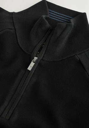 Črn zip-up pulover z visokim ovratnikom, ki ima rebrasto teksturo na ovratniku in manšetah, ter srebrno zadrgo kot poudarek.