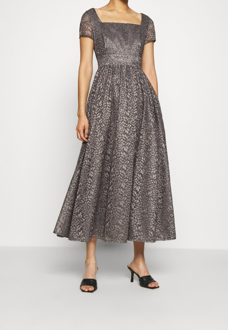 Femme portant une robe midi en dentelle grise avec manches courtes, encolure carrée, taille cintrée et sandales à talons noires ouvertes à l'avant.