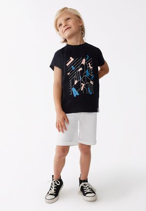 Camiseta negra de manga corta con diseño gráfico geométrico multicolor, combinada con pantalones cortos de mezclilla blancos y zapatillas negras.