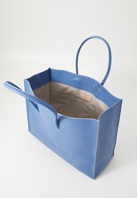 Sac fourre-tout en cuir bleu avec surface texturée, ouverture sur le haut, deux poignées et un intérieur beige avec une étiquette de marque.