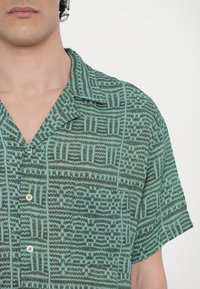 Camisa de botão de mangas curtas com um padrão abstrato verde e teal, design com colarinho, textura suave e detalhe de bolso frontal.