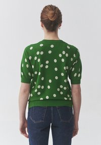 Pull court à manches courtes vert avec des pois blancs, col et poignets côtelés, texture douce, porté avec un jean bleu, vu de dos.