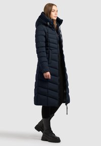 Marineblå pufferjakke med lang, tætsiddende design, hætte, sidelomme med lynlås og lodrette stikninger. Har en glat, blank overflade og snoretræk.