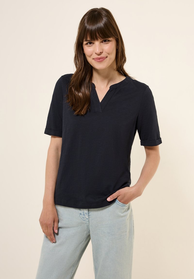 Haut bleu marine à manches courtes avec un col en V et des poignets retroussés, fabriqué dans un matériau doux. Porté avec un jean bleu clair, offrant une coupe décontractée.