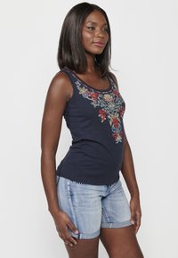 Marineblaues Tanktop mit buntem floralen Stickmuster am Ausschnitt, kombiniert mit hellblauen Denim-Shorts. Der Stoff scheint weich zu sein, das Design sitzt eng.