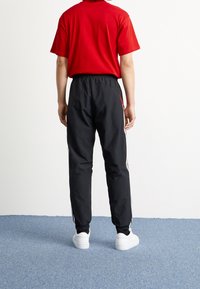 Röd t-shirt med kort ärm som matchas med svarta träningsbyxor med sidostriper och elastiska manschetter, bärs med vita sneakers på ett blått golv.