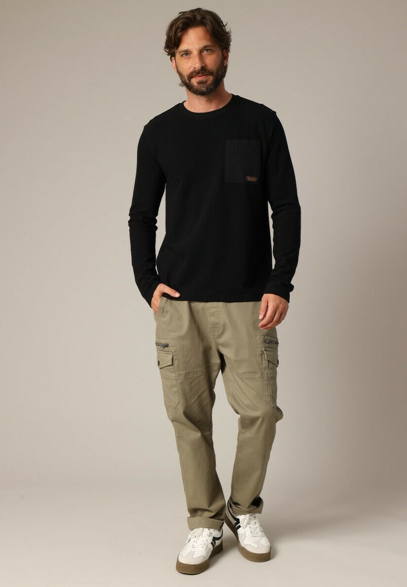 Chemise noire à manches longues avec une poche poitrine, associée à un pantalon cargo beige clair avec des poches zippées et des baskets blanches avec des accents noirs.