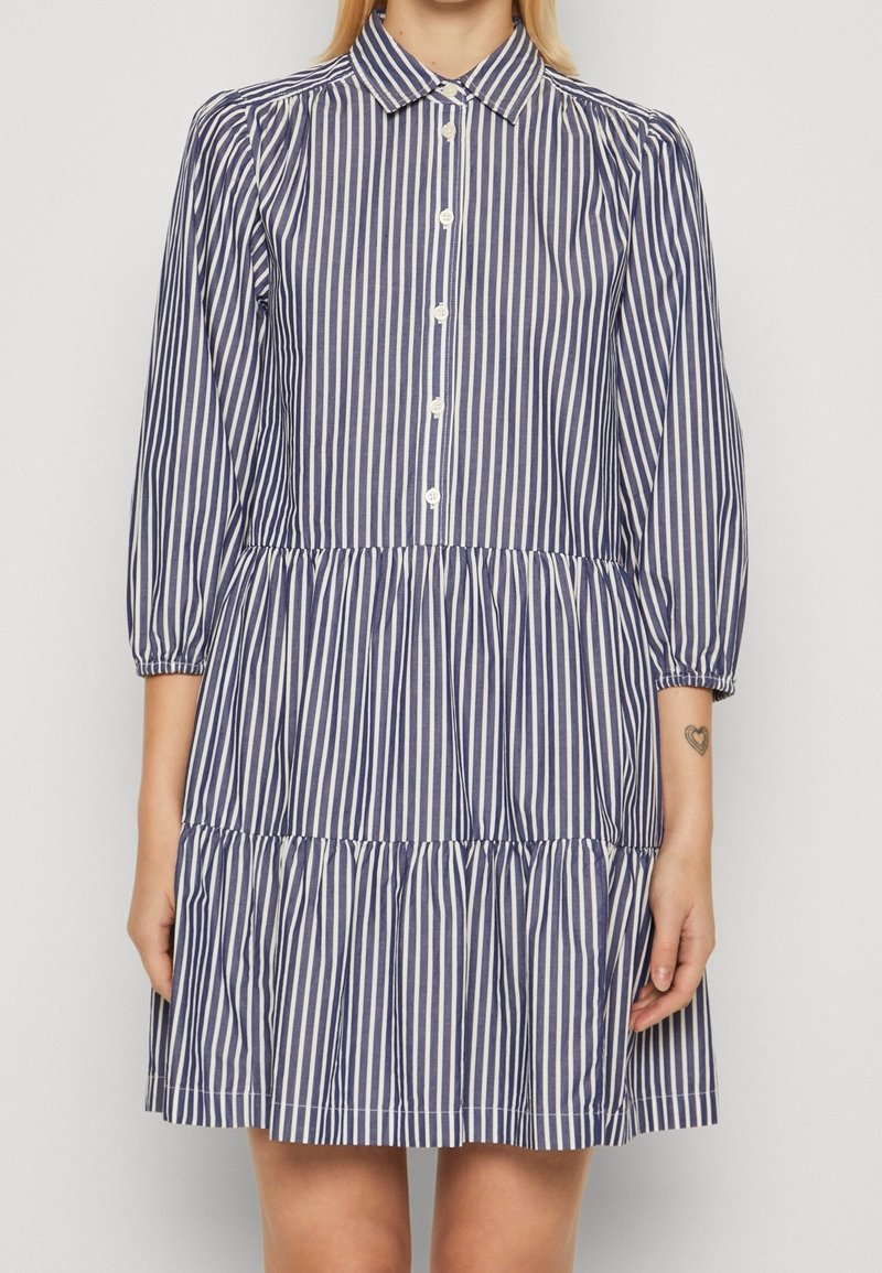 Robe rayée avec col, boutonnage sur le devant, manches 3/4 et jupe à volants. Les couleurs comprennent des rayures bleu marine et blanc.