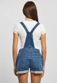 Denim overalls with adjustable straps, featuring a fitted bodice, rolled hem shorts, and two back pockets in a medium wash. White T-shirt underneath.