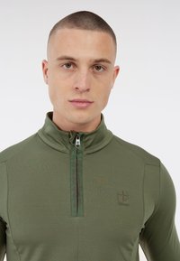 Primo piano di un uomo con capelli corti che indossa una felpa verde oliva con zip, con la scritta "PROTEST" nell'area della zip e il logo sul petto.