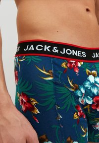 Rövid virágmintás boxer alsó, kék alapszínt visel, trópusi mintákkal piros, fehér és zöld színekben, fekete derékpánttal, amelyen a "JACK & JONES" felirat szerepel.
