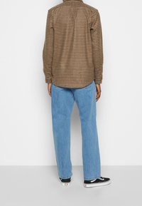 Ruskea ruudullinen flanellipaita, jossa on rento istuvuus, napitetut hihansuut ja selkäkaulus, yhdistettynä vaaleansinihin suoriin farkkuihin ja mustiin lenkkareihin.