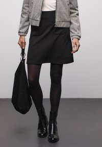 Černá mini sukně s texturou a dvěma předními kapsami, kombinovaná se šedou bomber bundou. Černé punčocháče a kotníkové boty doplňují outfit, drží černou tašku.