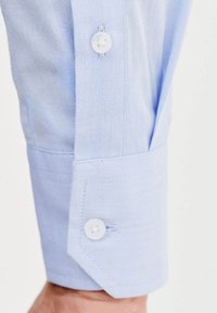 Jasnoniebieski mankiet koszuli z długim rękawem z teksturowanej tkaniny, wyposażony w zapięcie na guzik i skośny wzór na końcu rękawa.