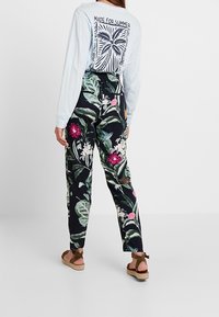 Svart byxor med blommönster pryds av färgglada blommor och löv, som kombineras med en ljusblå skjorta med långa ärmar. Modellen bär bruna sandaler med remmar.