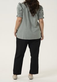 Lichtgroene blouse met opgezette korte mouwen, gladde stof, relaxte pasvorm; gecombineerd met zwarte broek en beige schoeisel.