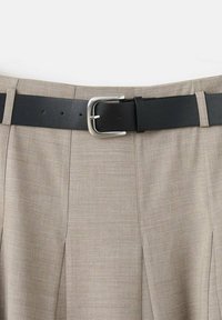 Pantalones beige plisados con un cinturón de cuero negro y una hebilla plateada, que destacan una textura suave y un diseño a medida.