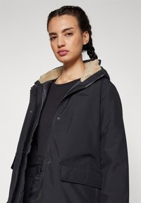 Veste extérieure noire avec une capuche en polaire, présentant un devant zippé, des poches latérales et une texture lisse. Logo sur la poitrine gauche.