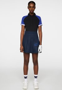 Svart pikétröja med blå raglanärmar, matchad med en marinblå plisserad kjol med blå akcentmönster. Vita golfhandskar och skor.