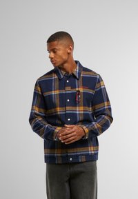 Marine rutete jakke med oransje og gule detaljer, laget av mykt stoff, med knappestolpe foran og en karabinert festet.