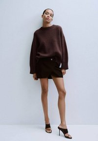 Hnědý pletený svetr, černá zavinovací sukně a černé pantofle na podpatku. Modelka stojí na světle šedém pozadí, aby ukázala střih a texturu outfitu.