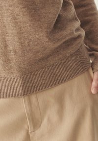 Pull en tricot marron avec ourlet côtelé, associé à un pantalon beige. Texture visible et matière douce et chaude mise en valeur dans une coupe décontractée.