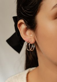 Srebrne kolczyki w formie kółek, z trzema połączonymi pierścieniami. Kolczyki są prezentowane na uchu modelki z ciemnymi włosami i czarną wstążką.