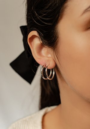 Silberne Creolen mit drei miteinander verbundenen Ringen. Die Ohrringe werden am Ohr eines Modells mit dunklem Haar und einem schwarzen Band präsentiert.