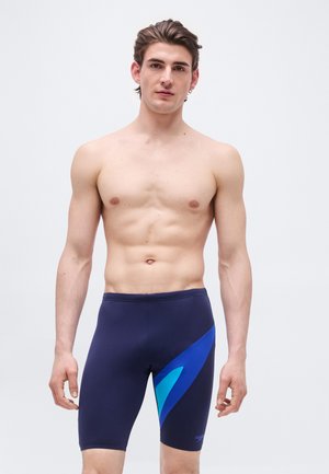 Nuori mies, jolla on märät hiukset ja joka pukeutunut tummansinisiin swim jammereihin, joissa on sinisiä yksityiskohtia, seisoo vaaleanharmaan taustan edessä.