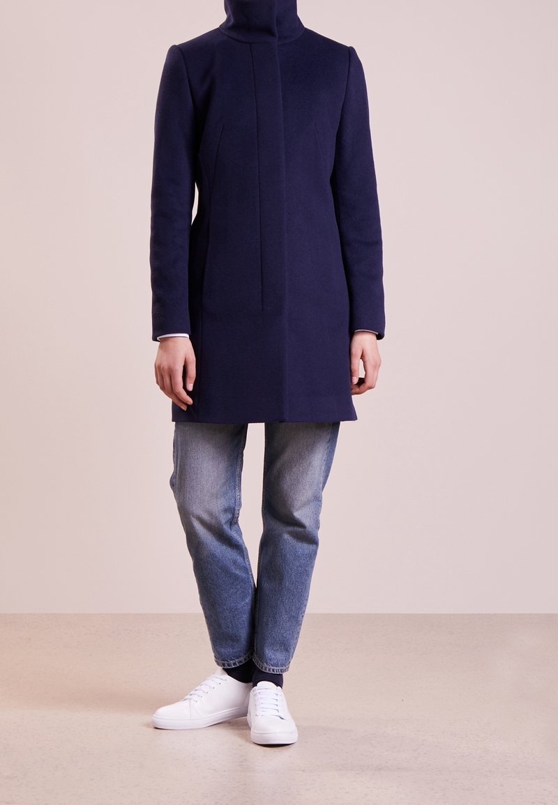 Navy wool coat with a high collar and vertical seam detailing, worn over light blue jeans and white sneakers on a neutral background.