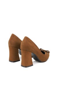 Scarpe con tacco alto in suede marrone, con punta arrotondata e un vistoso fiocco sul davanti; presentano un tacco spesso e una suola nera liscia.