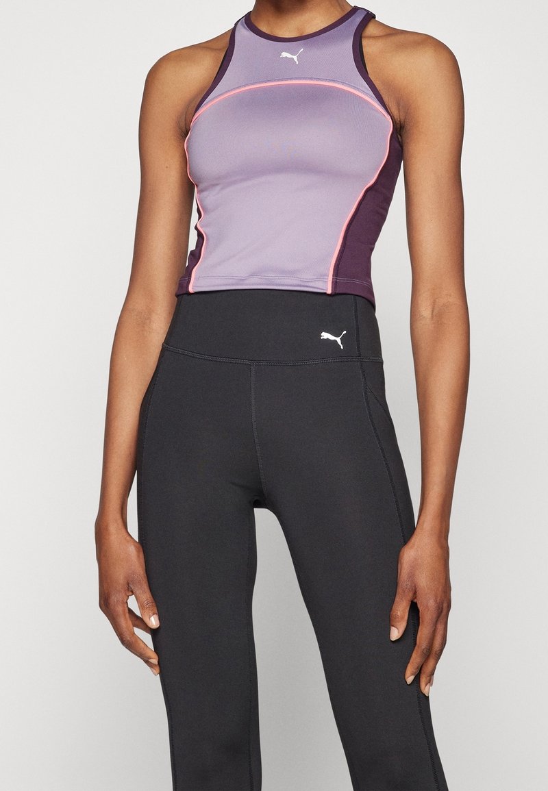 Donna che indossa un top sportivo senza maniche viola della Puma e leggings neri a vita alta sempre della Puma, in piedi con le braccia rilassate lungo i fianchi.