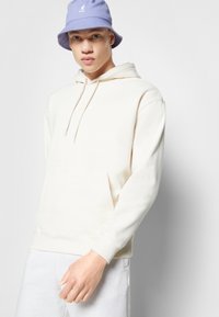 Haut à capuche beige clair avec une poche kangourou et un capuchon à cordon, associé à un chapeau seau violet clair comportant un logo. Texture lisse.