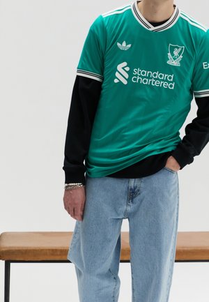Persoon draag een groen Liverpool voetbalshirt over zwarte lange mouwen en lichtblauwe jeans, staand bij een houten bankje tegen een effen achtergrond.