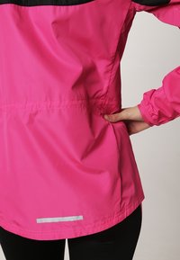 Personne portant une veste rose vif avec des bordures noires, la main placée dans la poche latérale, debout contre un fond clair uni.