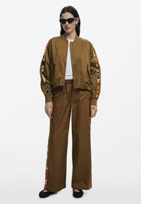 Hnedá bomber bunda s farebnými vzorovanými rukávmi, doplnená o zodpovedajúce hnedé nohavice so bočnými akcentmi. Hrubá tkanina, voľný strih.