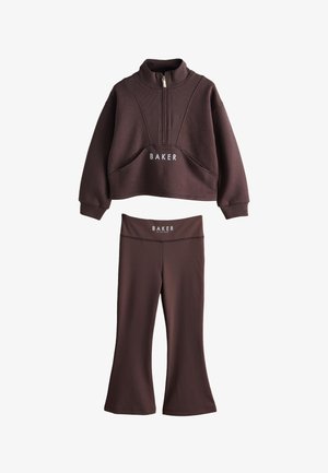 Blondynka zwrócona w lewo, ubrana w brązowy sweter z nadrukiem "BAKER BY TED BAKER" na plecach w pobliżu kołnierza.