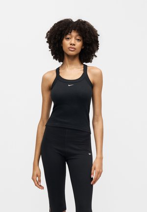 Vrouw met krullend haar in een zwarte Nike tanktop en zwarte legging, staand tegen een effen witte achtergrond.