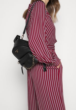 Mini-sac à dos en cuir noir à finition texturée, avec une sangle chaîne dorée en accent et une poignée plate sur le dessus, porté avec une tenue bordeaux à rayures.