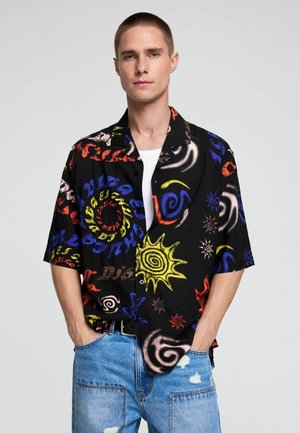 Mann mit kurzen Haaren trägt ein schwarzes Kurzarmshirt mit bunten abstrakten Sonnen- und Spiralmustern, kombiniert mit zerrissenen blauen Jeans.
