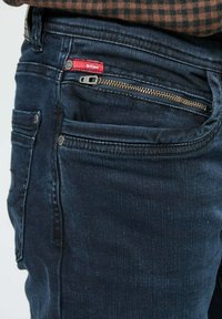 Jean bleu foncé avec une coupe slim, comportant une poche avant avec une fermeture éclair visible, des accents en métal et un patch de marque rouge.